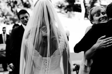 Braut in weißem Kleid mit langen Schleier, umgeben von Gästen bei einer Hochzeit.
