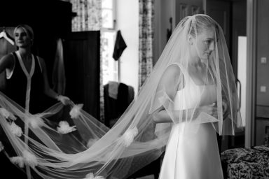 Eine Braut in einem weißen Kleid, die einen langen Schleier trägt, wird von einer Frau unterstützt.