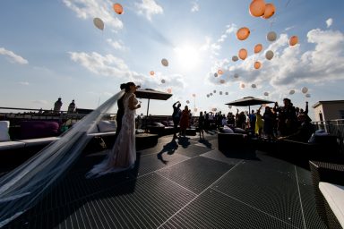 Braut in Hochzeitskleid, umgeben von Gästen und schwebenden Ballons bei Sonnenlicht.