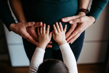 Hände um einen runden Bauch, ein Kind berührt spielerisch die Hände der Erwachsenen.
