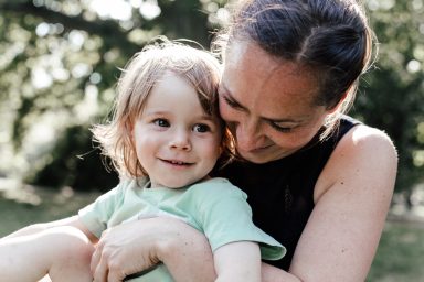 Eine lächelnde Mutter hält ein Kind im Freien, umgeben von Natur.