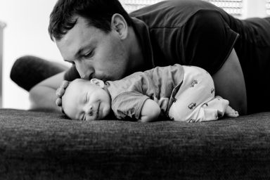 Vater küsst lächelnd sein schlafendes Baby auf einer Couch.