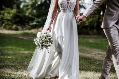 Braut in weißem Kleid mit Bouquet, Hand in Hand mit Bräutigam im Freien.