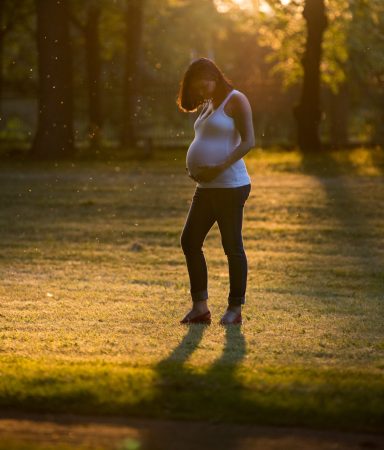 Schwangere Frau steht im Sonnenuntergang auf einer Wiese, umgeben von Bäumen.