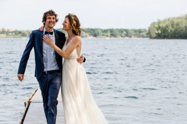 Braut und Bräutigam stehen am Wasser und lachen, umgeben von einer malerischen Landschaft.